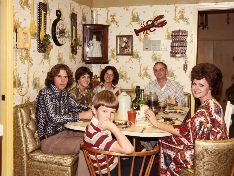 Six people sit around a small dining table eating a meal in a vintage kitchen with patterned wallpaper, gold chairs, and 1970s decor including a rotary phone and lobster wall hanging. Everyone is looking toward the camera.