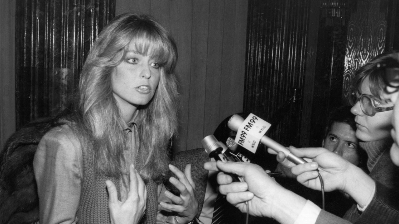A woman with long hair speaks animatedly to reporters holding microphones toward her, while photographers and another person look on. The image is black and white and appears to be from the 1970s or 1980s.