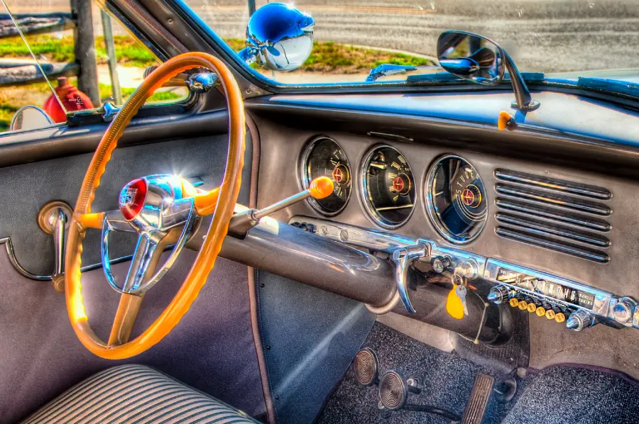 Close-up of a vintage car’s interior showing a striped seat, wooden steering wheel, classic gauges, radio, and yellow key on the dashboard. Bright sunlight highlights the retro details.