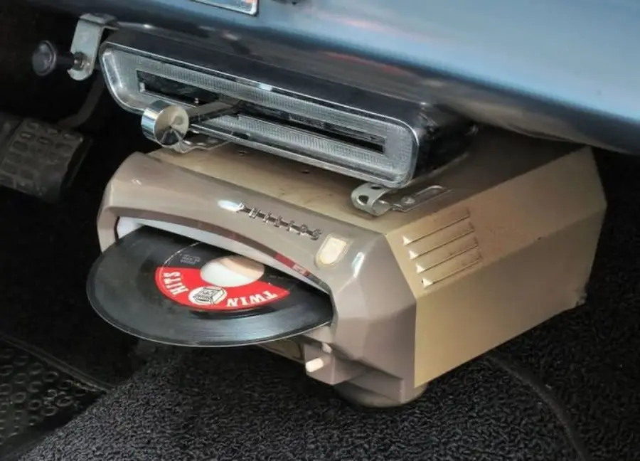A vintage car dashboard with a built-in record player under the radio, playing a 45 rpm vinyl record. The car’s pedals and carpeted floor are visible in the background.