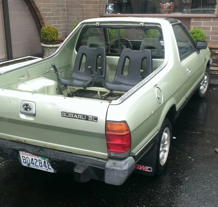 A light green Subaru GL Brat with two rear-facing seats in the truck bed, parked outside on a wet surface near a brick building and plants.