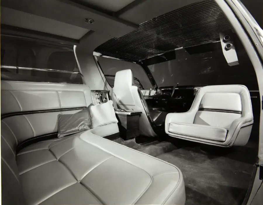 Futuristic car interior with white leather seats, a curved sofa in the back, and a swivel front passenger seat. The dashboard is sleek, with a horizontal bar dividing the cabin. The overall design is spacious and modern.