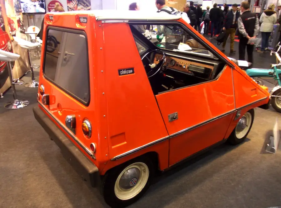 A small, red, three-wheeled car with an angular design is displayed indoors. People are visible in the background at what appears to be a car show or exhibition.