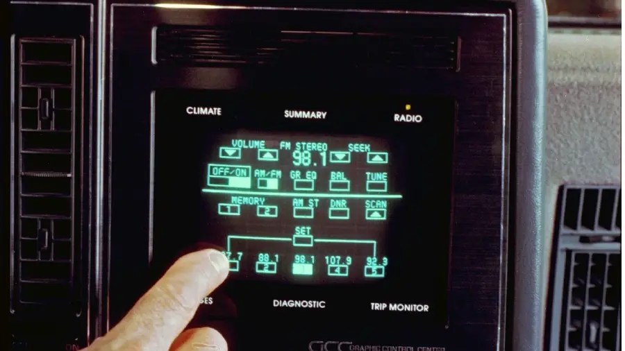 A finger touches the touchscreen radio controls on a retro digital car dashboard display. The screen shows FM radio settings, volume, and memory presets in green text.