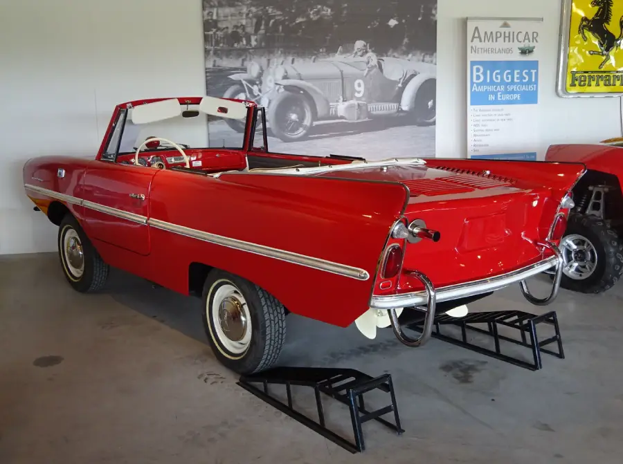 A red vintage Amphicar convertible is displayed indoors with its rear wheels elevated on ramps. Classic car posters and a Ferrari logo are visible on the wall behind it.