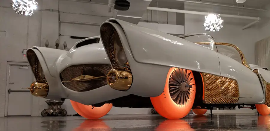 A futuristic white and gold car with glowing orange wheels is displayed indoors under bright lights, giving the vehicle a unique, retro-futuristic look.