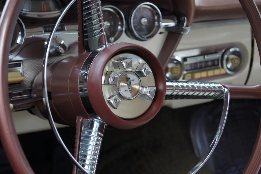 Close-up of a classic car steering wheel with chrome accents, vintage dials, and a dashboard featuring analog gauges and buttons.