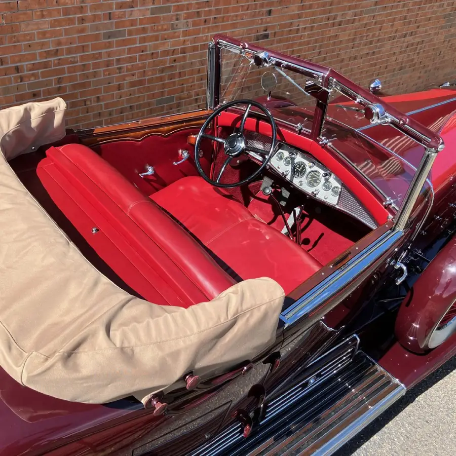 A vintage convertible car with red interior, chrome details, and a tan folded soft top is parked next to a brick wall. The dashboard features classic gauges and a large steering wheel.