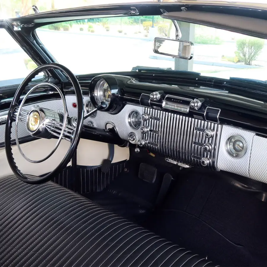 Interior of a vintage car showing a large steering wheel, classic dashboard with chrome details, analog dials, buttons, a radio, and black-striped bench seat upholstery.