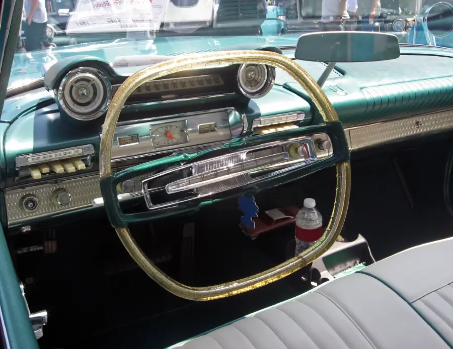 Classic car interior with a unique gold-colored square steering wheel, vintage dashboard, analog clock, radio, and a plastic water bottle on the seat. Sunlight highlights the teal and green colors.