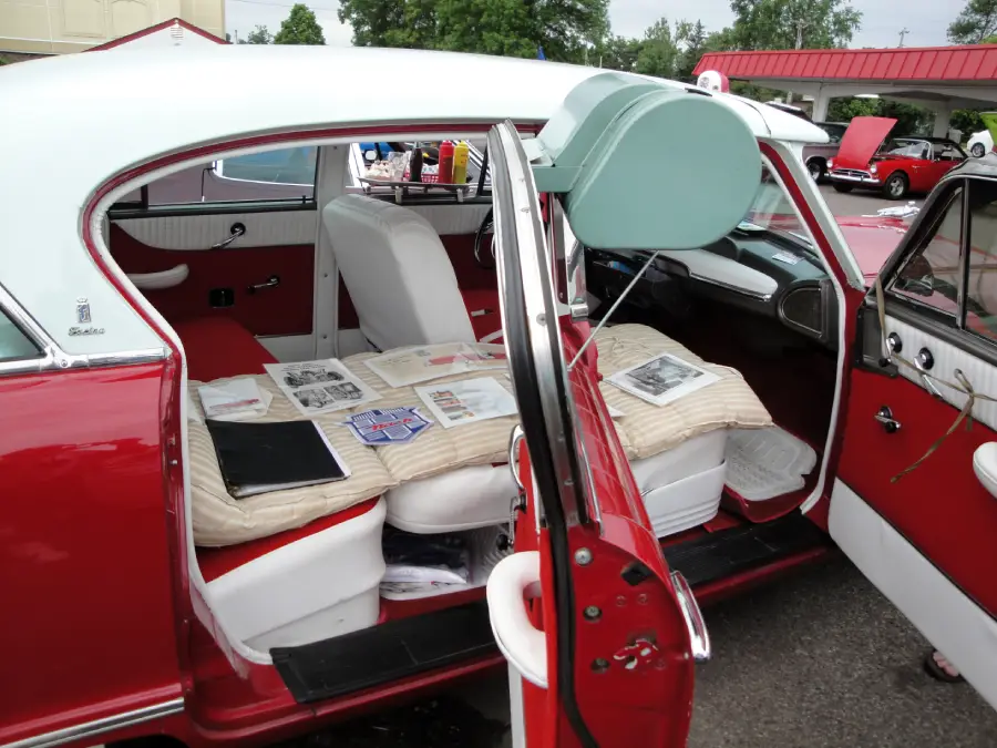 A classic red and white car with open doors, showing white seats and vintage documents displayed on a beige blanket across the back seat. Other cars and people are visible in the background at an outdoor event.