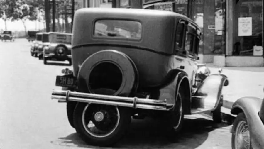 Black-and-white photo of a vintage car parked on a city street, viewed from the rear. Other classic cars and storefronts are visible in the background.