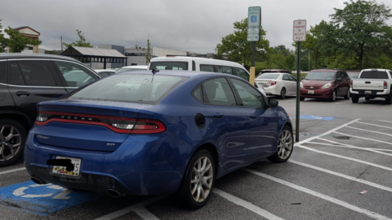 A blue Dodge sedan is parked over two handicapped parking spaces on a cloudy day. Other vehicles are properly parked nearby. The area looks like a public parking lot with trees and buildings in the background.