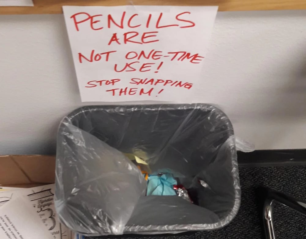 A trash can sits below a sign that reads, “PENCILS ARE NOT ONE-TIME USE! STOP SNAPPING THEM!” The trash contains crumpled papers and waste.