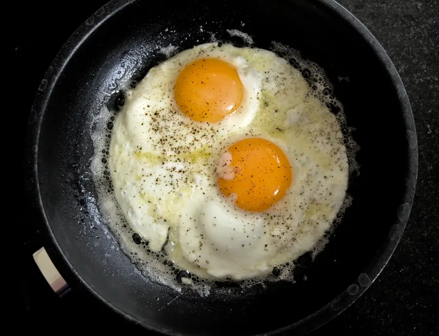 Two sunny-side-up eggs cooking in a black frying pan, with salt and pepper sprinkled on top. The egg whites are set, and the yolks are still runny. The pan is on a dark surface.