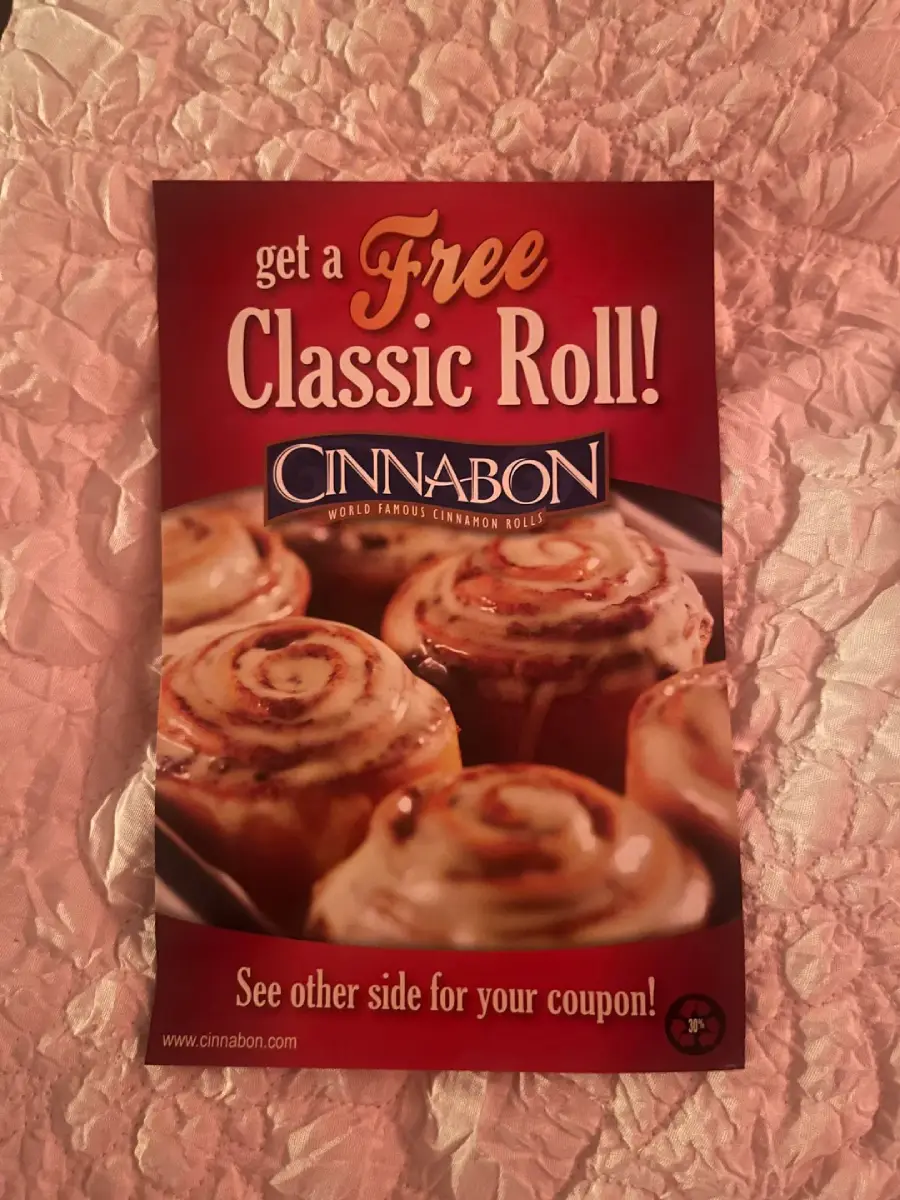 A Cinnabon flyer on a pink textured surface offers a free Classic Roll, showing iced cinnamon rolls and the Cinnabon logo. Text at the bottom mentions a coupon on the other side.