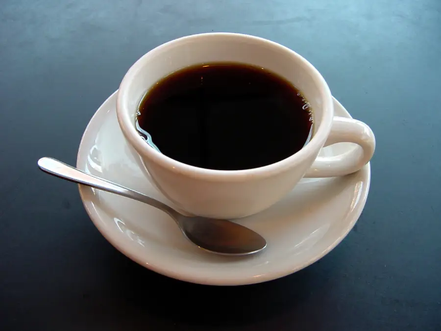 A white cup filled with black coffee sits on a matching saucer, with a silver spoon resting on the saucer, all placed on a dark surface.