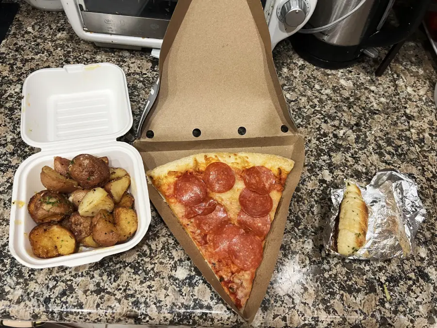 A large slice of pepperoni pizza in a pizza box, roasted potatoes in a white takeout container, and a piece of foil-wrapped garlic bread on a granite countertop.