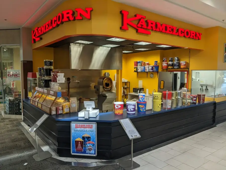 A Karmelcorn store in a mall features a yellow counter, popcorn machines, various popcorn tins on display, and a red Karmelcorn sign above. A GNC store is visible to the left.