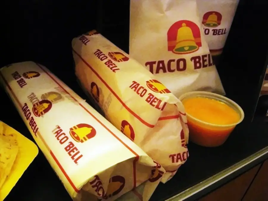 Two wrapped Taco Bell burritos, a bag with the Taco Bell logo, a small container of nacho cheese sauce, and part of a taco or quesadilla are visible on a black surface.