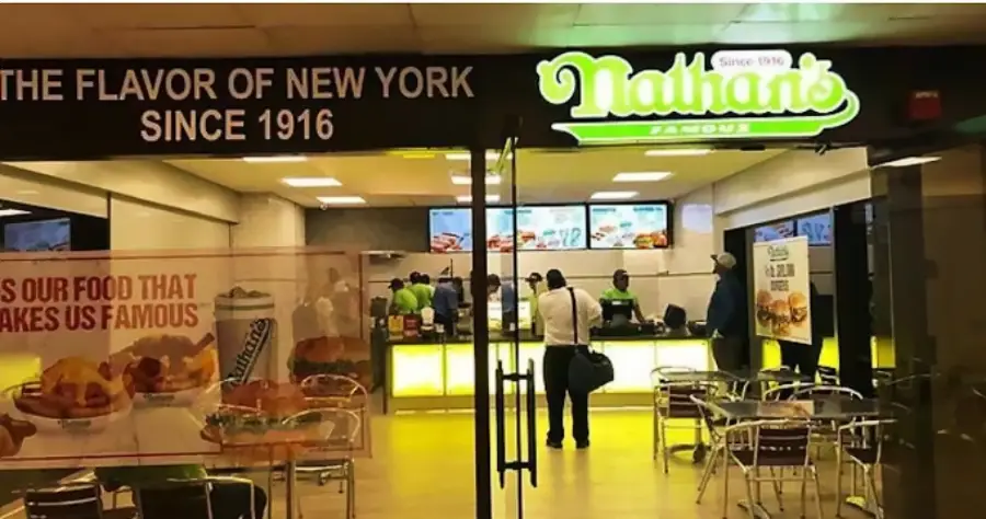 Nathan’s Famous restaurant with neon green signage, slogan “The flavor of New York since 1916,” visible food posters, menu screens, glass doors, and a few people ordering at the counter. Tables and chairs in the foreground.