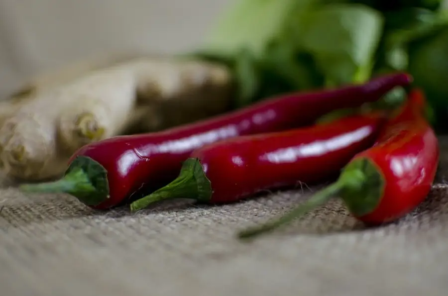Three red chili peppers with green stems rest on a burlap surface, with blurred ginger root and leafy greens in the background.