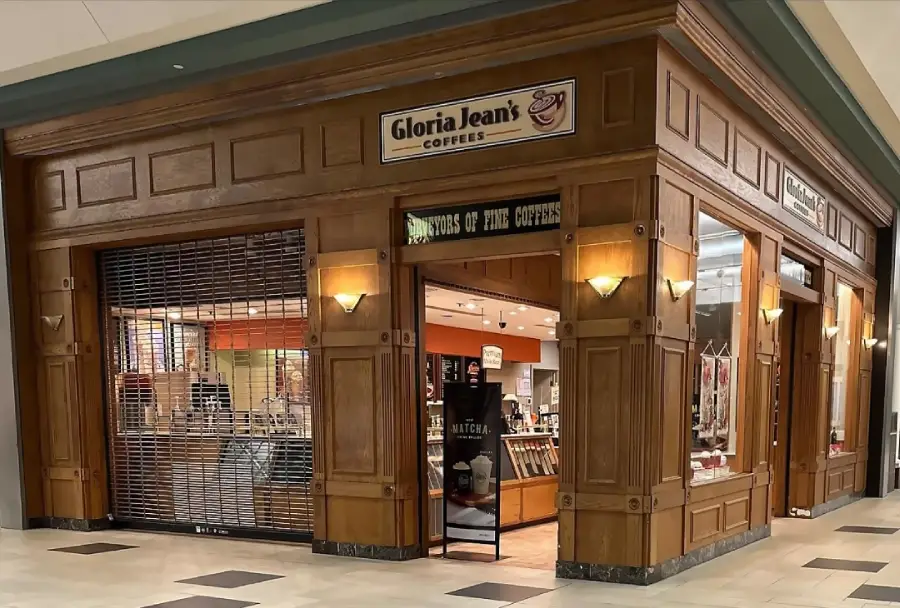 A Gloria Jean’s Coffees shop in a mall features wooden paneling, wall lamps, and a partially closed security gate. The interior is visible, showing shelves and a counter with coffee products.