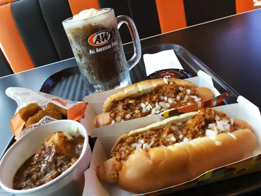 A&W meal with two chili cheese dogs topped with onions, a cup of root beer, a bowl of chili with cheese, and a side of fried cheese curds on a black tray.