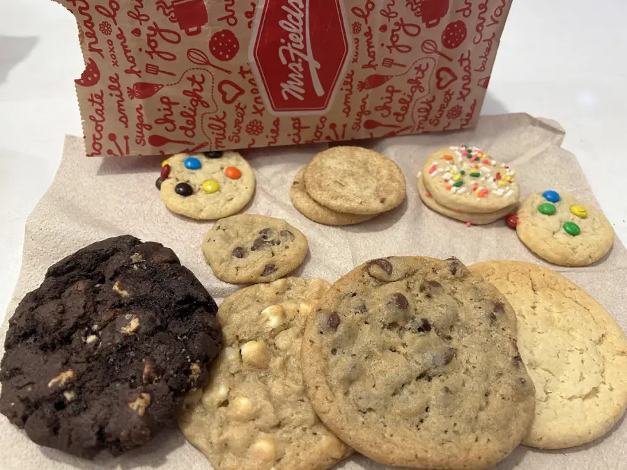 A variety of cookies, including chocolate chip, white chocolate, sprinkle, and M&M topped, are displayed on a napkin in front of a partially open Mrs. Fields box.