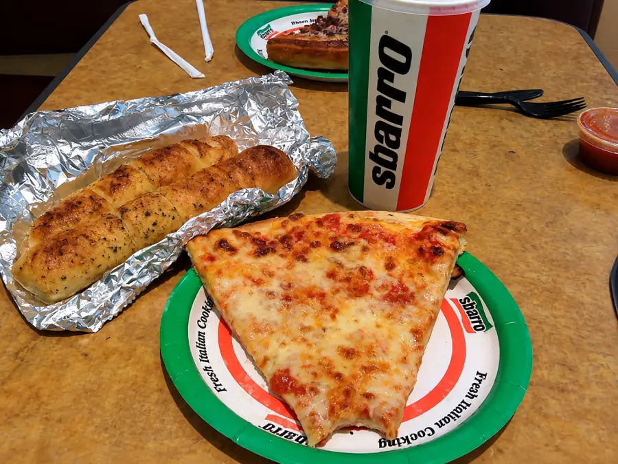 A slice of cheese pizza, garlic breadsticks in foil, and a large Sbarro drink cup sit on a table. Another pizza slice and a fork are visible in the background.