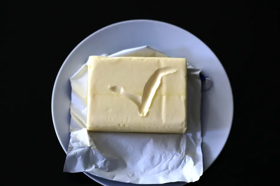 A rectangular block of butter with a “V” shaped mark on top, partially unwrapped, sits on a white plate against a black background.