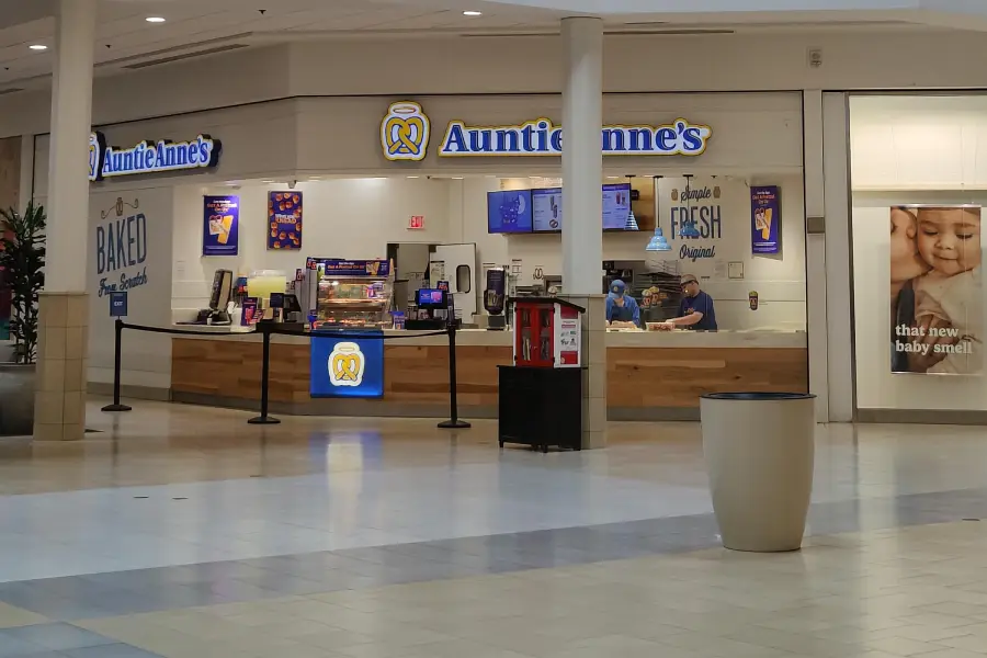 Auntie Anne’s pretzel shop in a mall, with staff working behind the counter. A sign and pretzel images are visible, along with nearby promotional posters and a trash can in the foreground.
