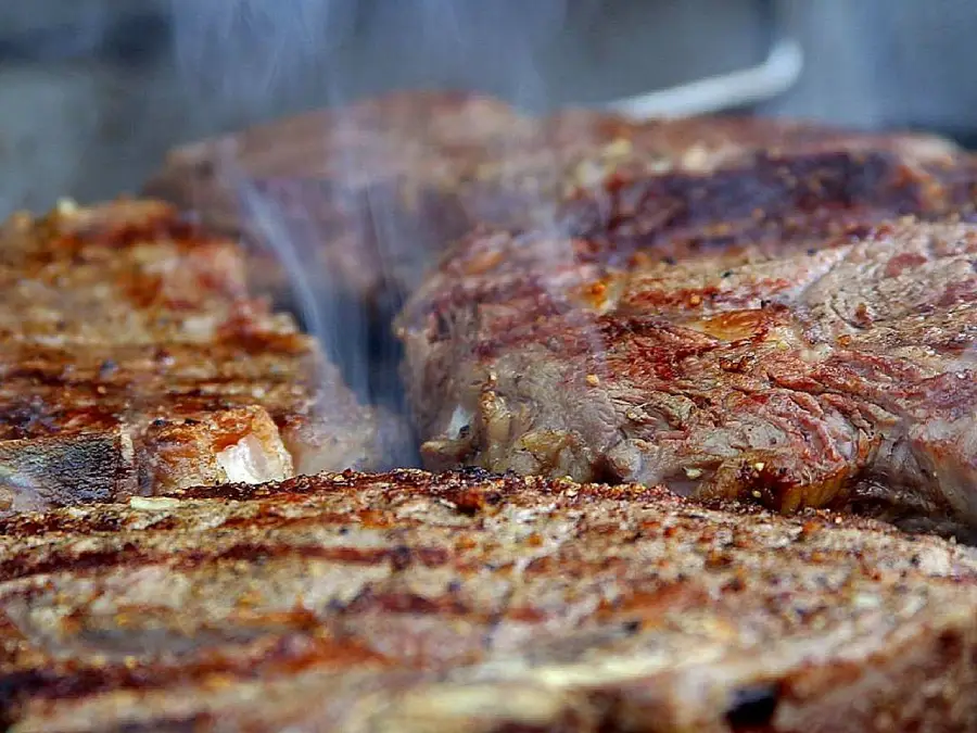 Close-up of several juicy, grilled steaks with visible grill marks, sizzling on a barbecue with smoke rising, highlighting the texture and doneness of the meat.