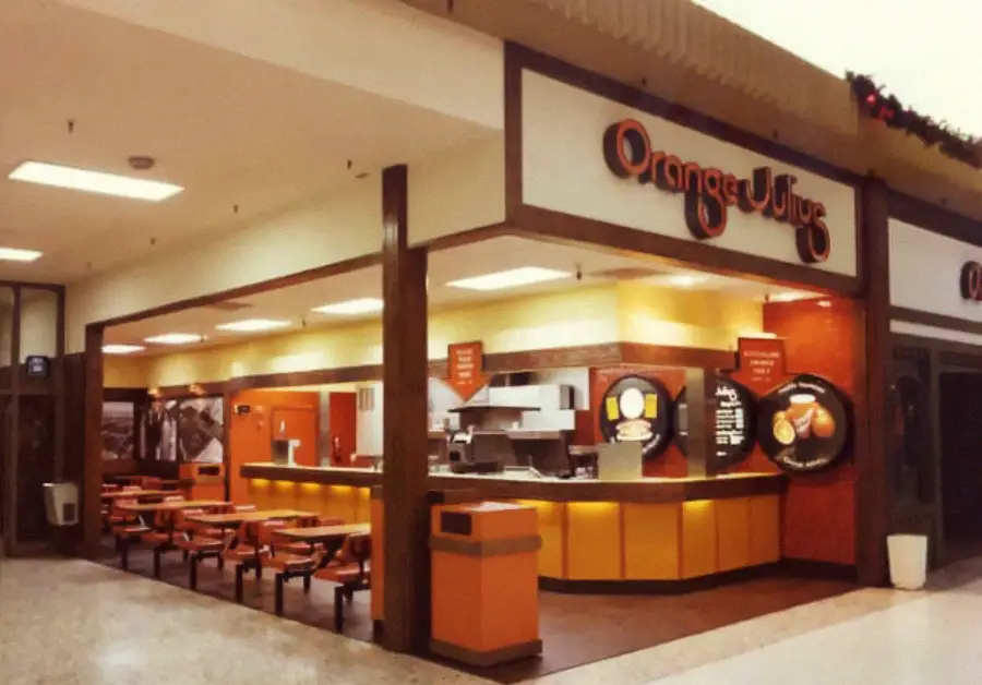 A vintage Orange Julius store in a mall, featuring orange and yellow counters, small tables with benches, and retro signage with the store’s name. The setting is brightly lit with a classic 1970s-1980s design.