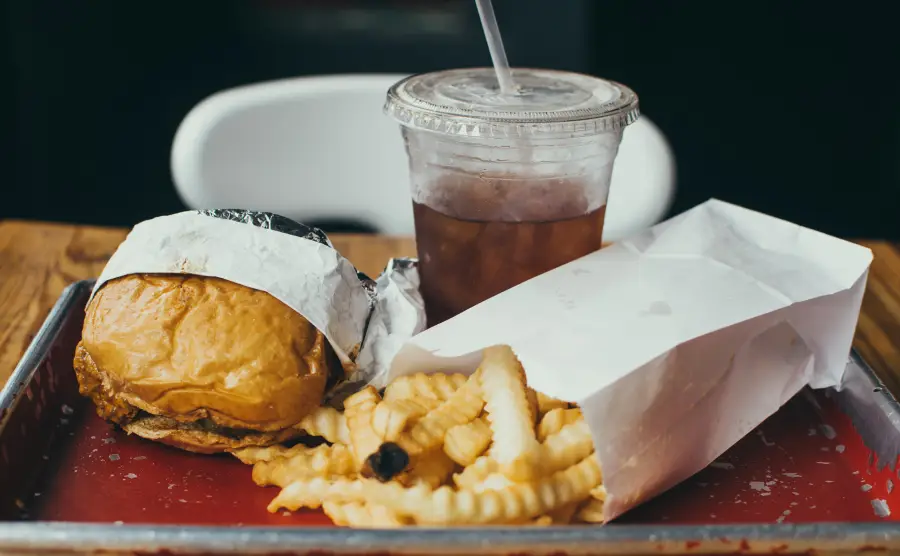 A tray with a wrapped burger, crinkle-cut fries in a paper bag, and a plastic cup of iced drink with a straw, set on a wooden table.