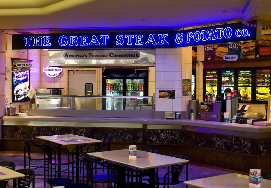 A food court restaurant called “The Great Steak & Potato Co.” features a lit sign, counter, menu boards, and empty tables with chairs in the foreground.