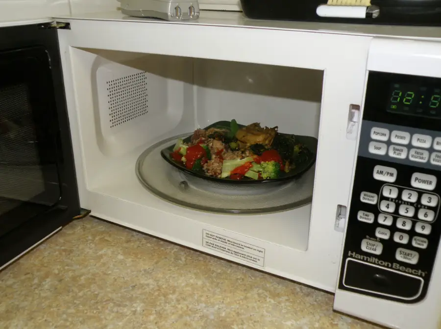 A plate of food with vegetables and grains sits inside a white microwave on a glass turntable. The microwave is on a beige countertop, and part of the control panel is visible.