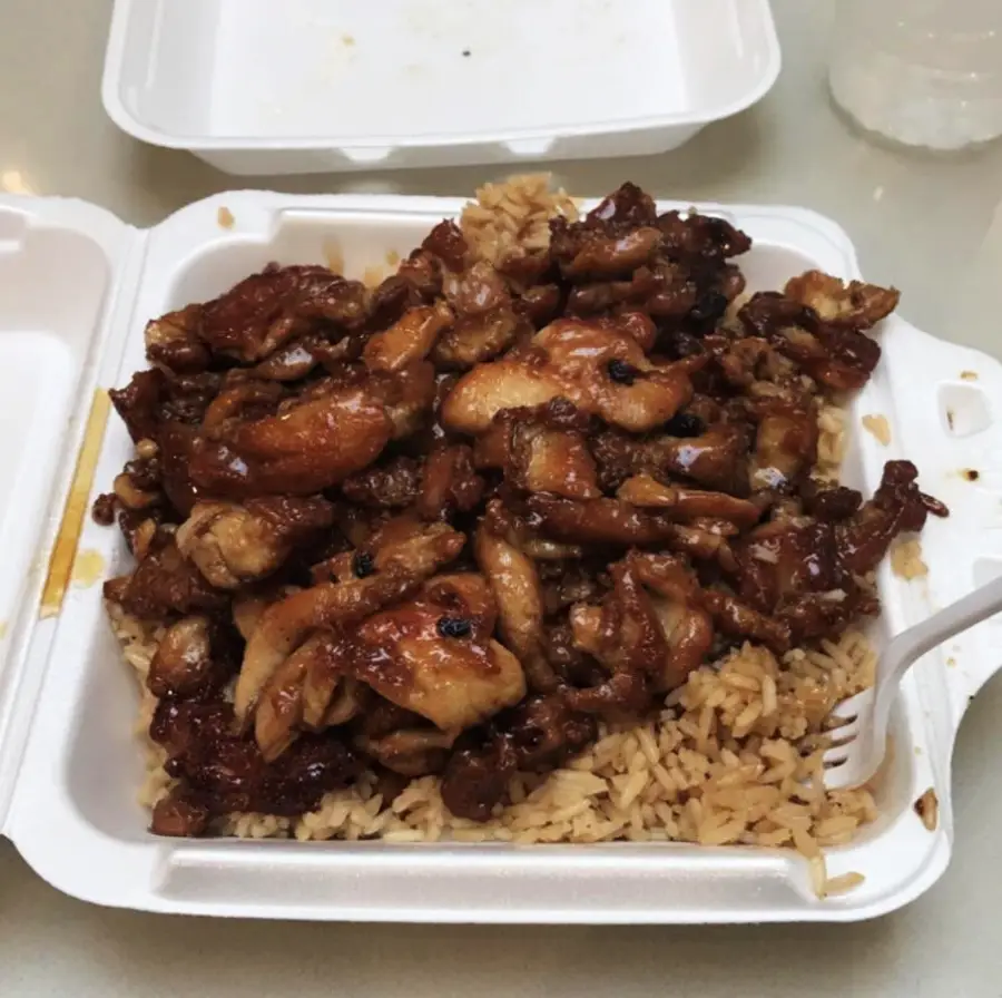 A white takeout container filled with grilled chicken pieces in a dark sauce, served over a bed of fried rice, with a white plastic fork on the side.