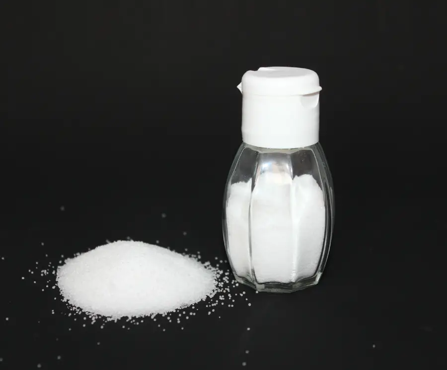 A small glass salt shaker filled with white salt sits beside a small pile of loose salt on a black surface, against a black background.