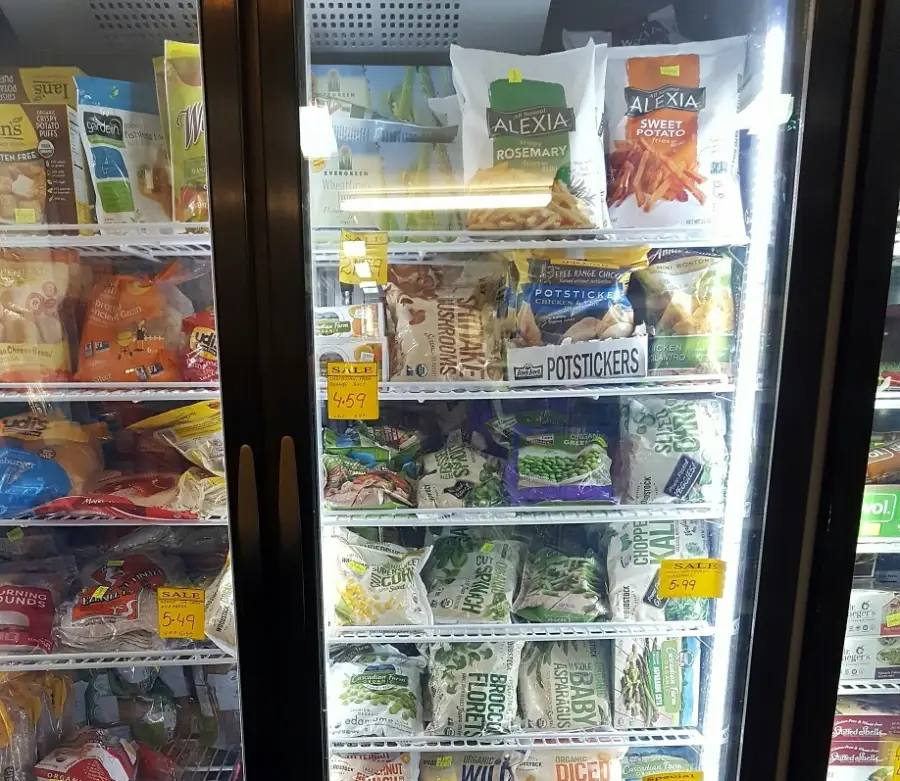 Freezer section in a grocery store with various frozen foods, including bags of Alexia fries, potstickers, and vegetables. Sale tags are visible on several shelves behind the glass doors.
