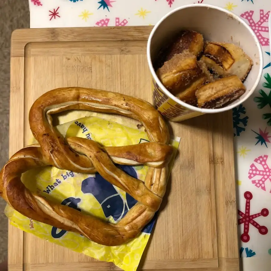 A large pretzel on a yellow wrapper and a cup filled with cinnamon sugar pretzel bites sit on a wooden cutting board, with a colorful festive tablecloth beneath.