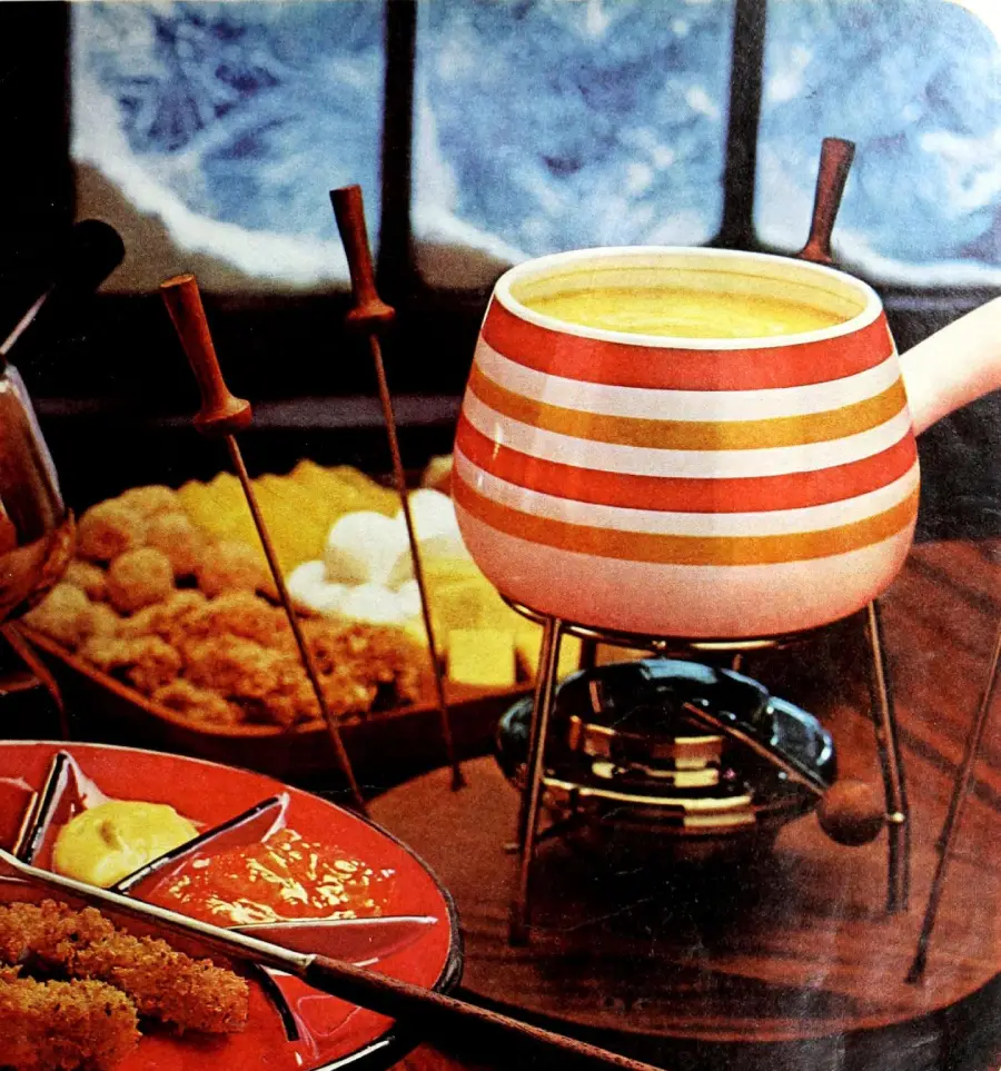 A striped fondue pot filled with cheese sits on a burner, surrounded by fondue forks, bread, vegetables, and a divided dish with dipping sauces. A snowy scene is visible through the window in the background.