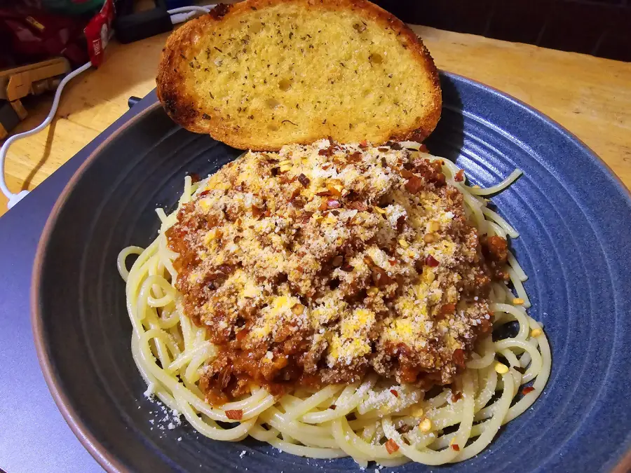 A plate of spaghetti topped with meat sauce and grated cheese, served with a slice of toasted garlic bread on the side.