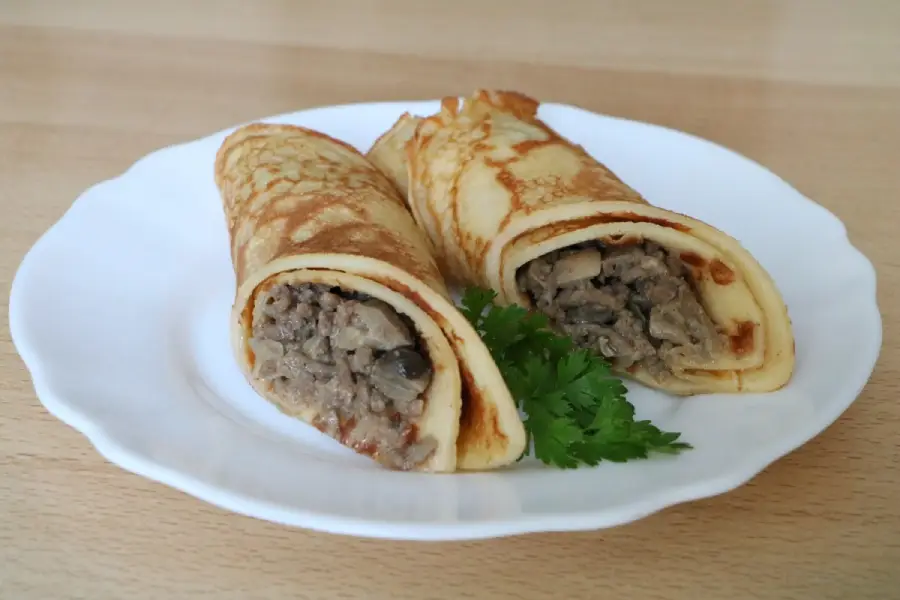 Two rolled crepes filled with a creamy mushroom and meat mixture, served on a white plate with a sprig of fresh parsley.