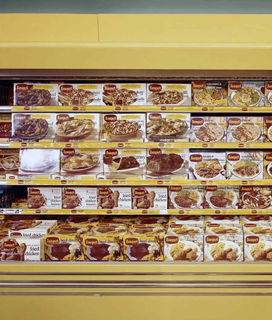 Frozen dinner boxes are stacked neatly on grocery store shelves, mostly yellow and white packaging, displaying various entrees such as fried chicken and meatloaf. The boxes are organized in rows inside a freezer section.