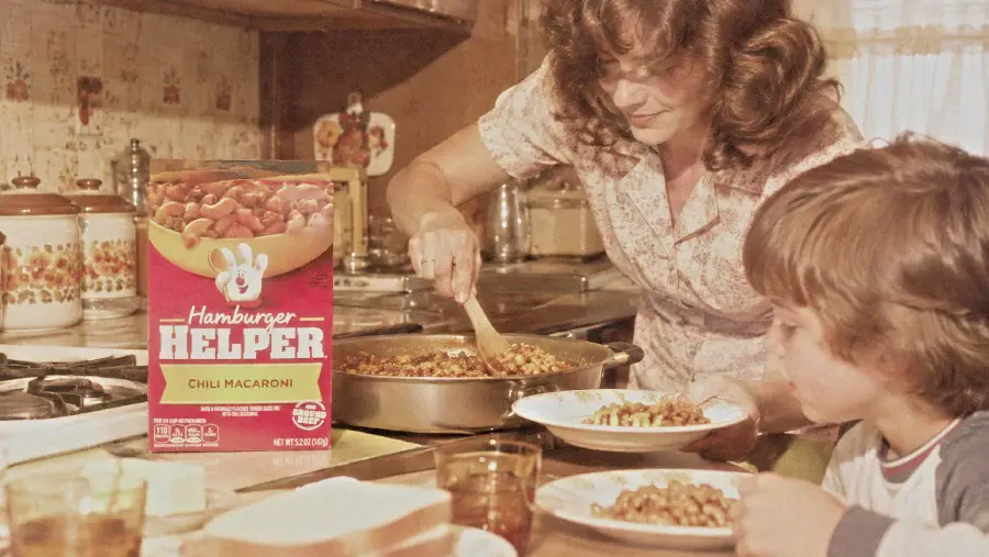 A woman serves Hamburger Helper chili macaroni from a skillet onto a boy’s plate at a kitchen table. The box is visible near the stove. Plates, glasses, and bread are on the table.