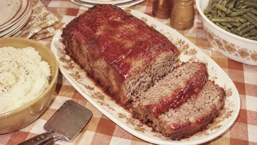 A platter with sliced meatloaf glazed with ketchup sits on a checkered tablecloth, next to a bowl of mashed potatoes, green beans, plates, and salt and pepper shakers.