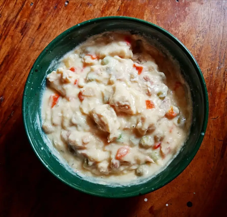 A green bowl filled with creamy chicken pot pie filling, containing chunks of chicken, peas, carrots, and sauce, sits on a wooden surface.