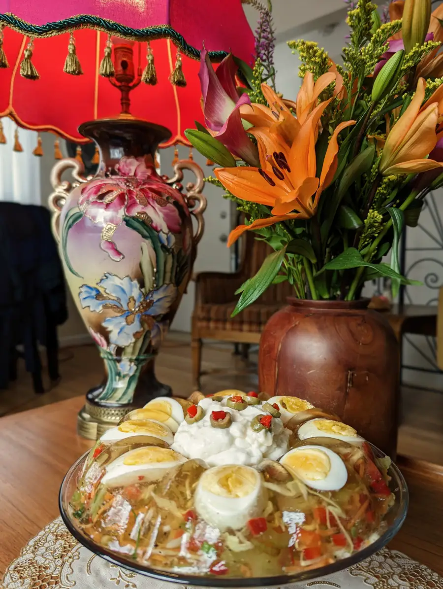 A decorative table with a colorful jelly salad topped with sliced eggs, a vintage floral lamp, and a vase of vibrant orange lilies.