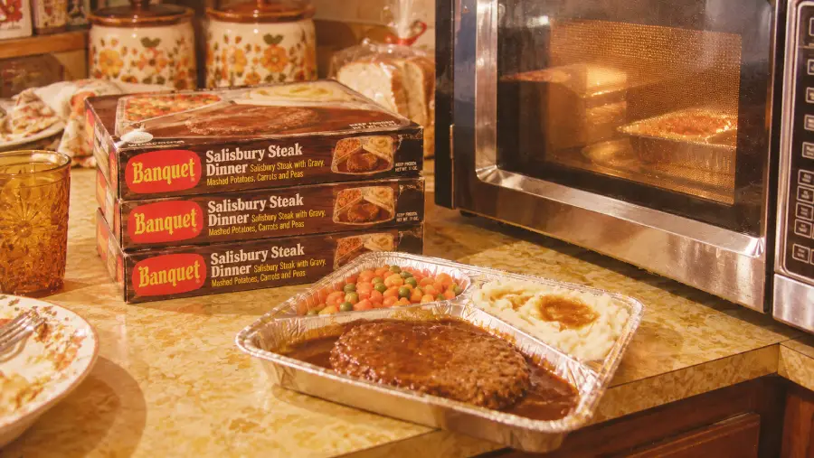 A Banquet Salisbury Steak frozen dinner sits on a counter next to a microwave, with two more dinner boxes stacked behind it. The meal includes steak, mashed potatoes, gravy, and mixed vegetables.